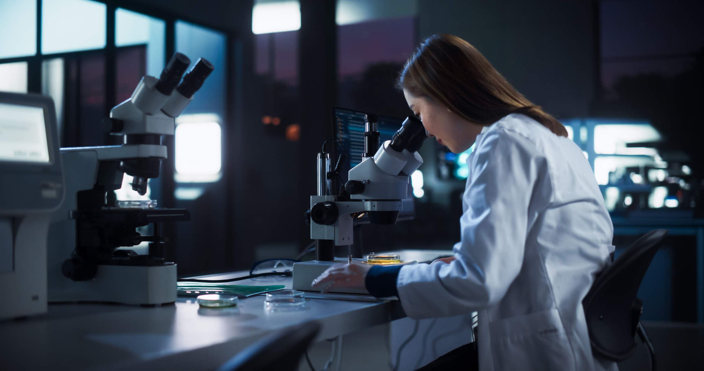 Medical Research and Development Laboratory: Hard-Working Asian Female Scientist Using Computer and Looking at a Sample in a Petri Dish Under a Microscope. Advanced Genetic Engineering Lab at Night